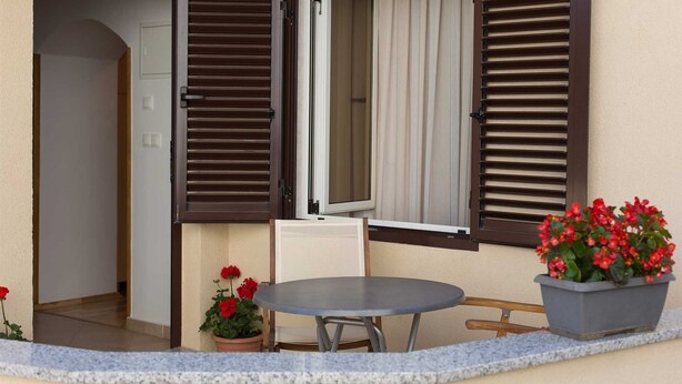 Un balcón abierto con una mesa y sillas, una maceta con flores rojas, y vista a un edificio a través de la puerta abierta.