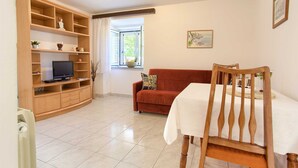 Una sala de estar con televisor, estanterías de madera, un sofá rojo, una mesa de comedor con una silla y una ventana con persianas.