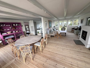 A spacious room with a wooden floor, a round dining table surrounded by wicker chairs, a purple bookshelf, and a fireplace. - Welcoming holiday home right down to the water (Rørvig)