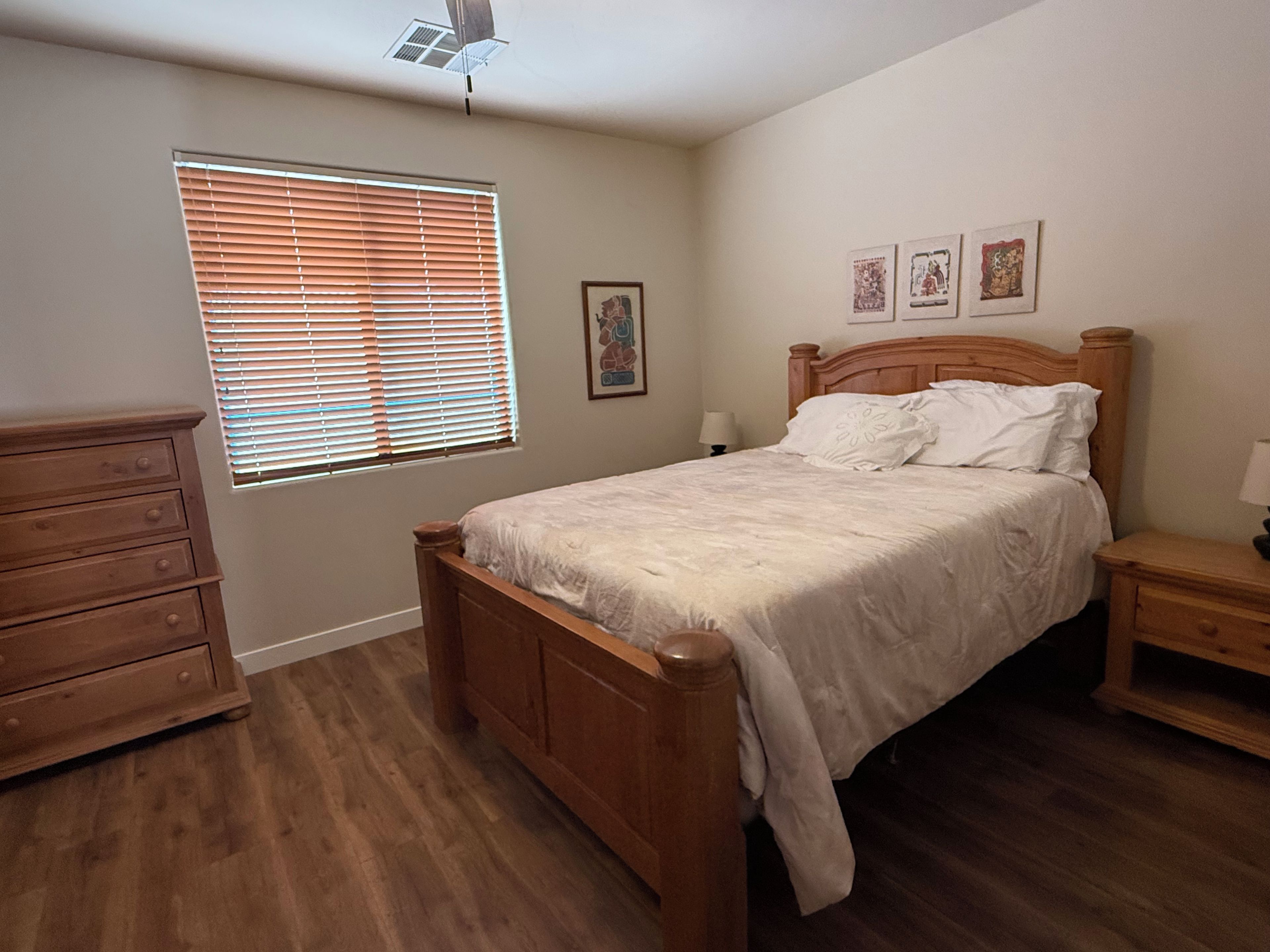 A bedroom with a wooden bed, a dresser, a window with blinds, and a wall with three framed pictures.