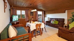 Ein gemütliches Blockhauss-Interieur mit Esstisch, Holzmöbeln, einem Fernseher und einer Couch.