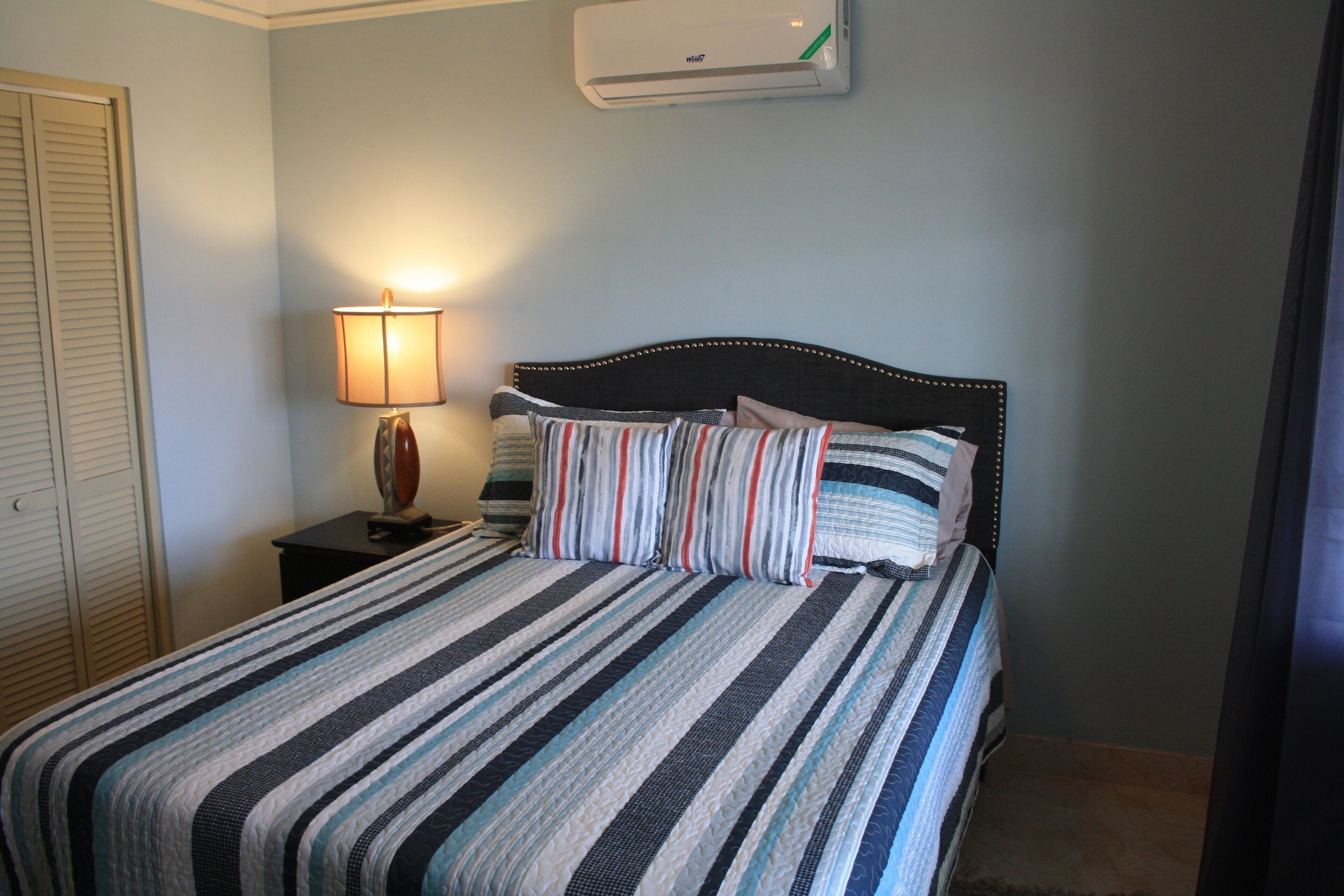 A neatly made bed with striped bedding, a bedside lamp, and an air conditioning unit on the wall.