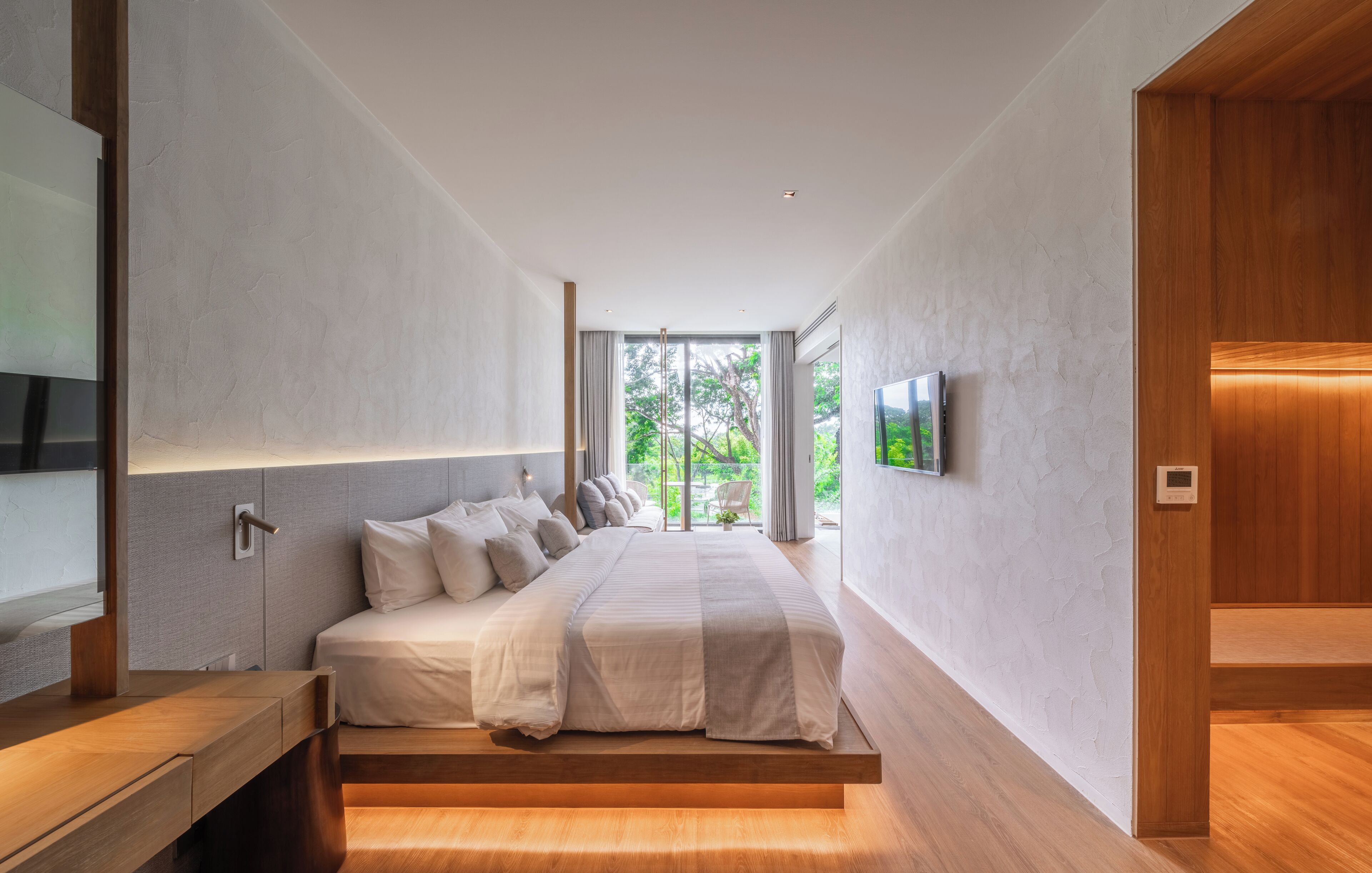 A modern bedroom with a large bed, built-in TV, and a view of greenery outside.