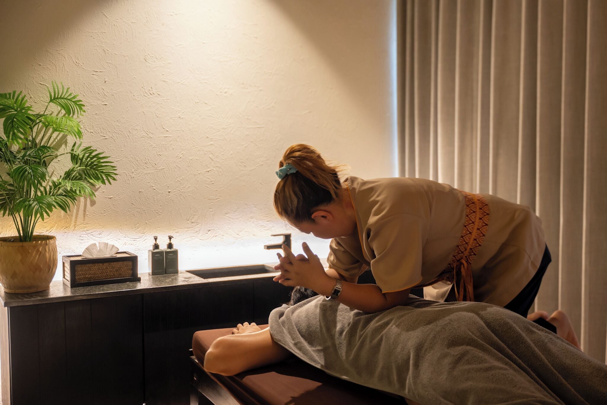 A person receiving a massage in a room with a plant, a tissue box, and a small bottle on the counter.