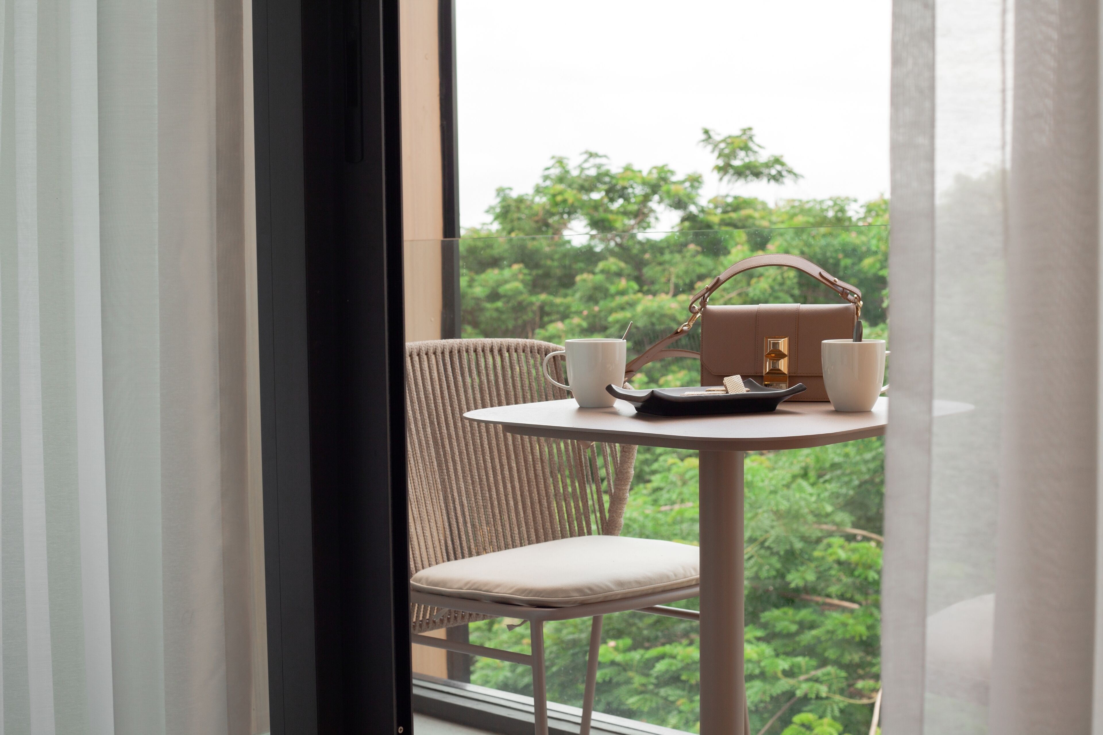 Una habitación con vistas a zonas verdes a través de una ventana, una mesa redonda con un bolso, dos tazas y una bandeja.