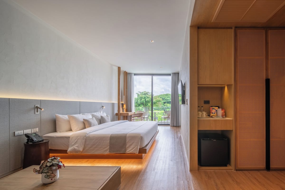 A modern hotel room with a large bed, bedside table, and a small desk. There is a view of greenery outside the window.