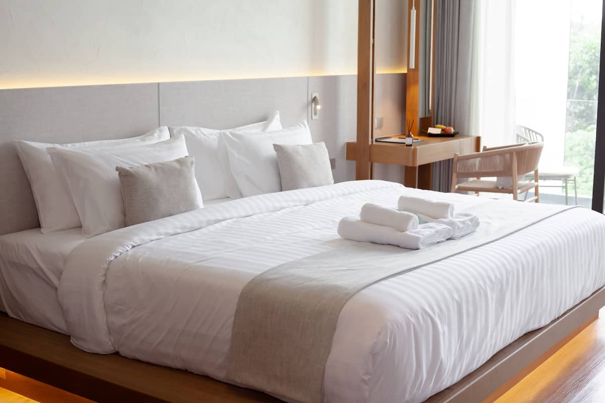 A neatly made bed with white linens and a grey throw blanket, a wooden nightstand with a lamp, and a view of greenery through the window.