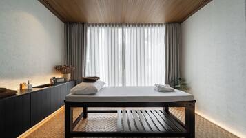A modern spa room with a wooden ceiling, a massage table, a sink with toiletries, and a large window with curtains.