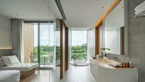 A modern bathroom with a large mirror, a bathtub, and a sink. There is a balcony with a view of trees.