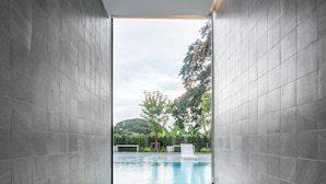 A modern bathroom with a glass-enclosed jacuzzi, grey tiled walls, and a view of an outdoor pool and greenery. - Riva Vista Riverfront Resort Chiangrai (Chiang Rai)