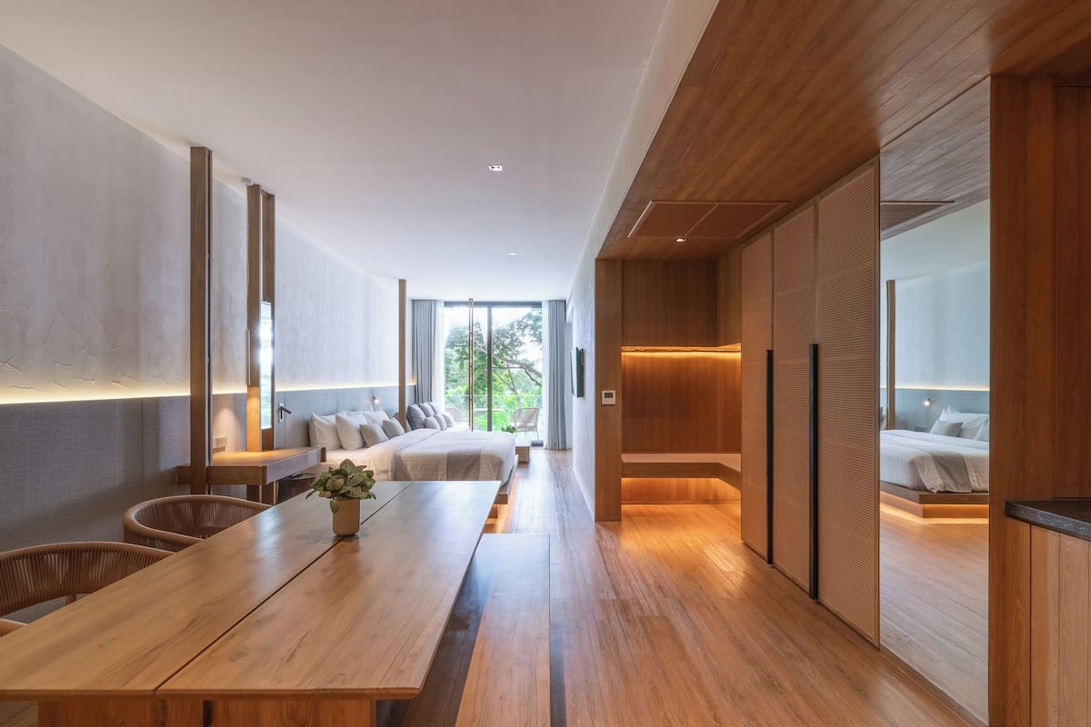 A modern hotel room with a wooden dining table, a sofa, and a bed with a view of the outdoors.