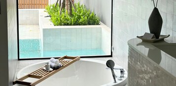 Casa de banho moderna com banheira, bandeja de madeira com toalhas e uma chávena, e uma janela ampla com vista para uma piscina e vegetação.