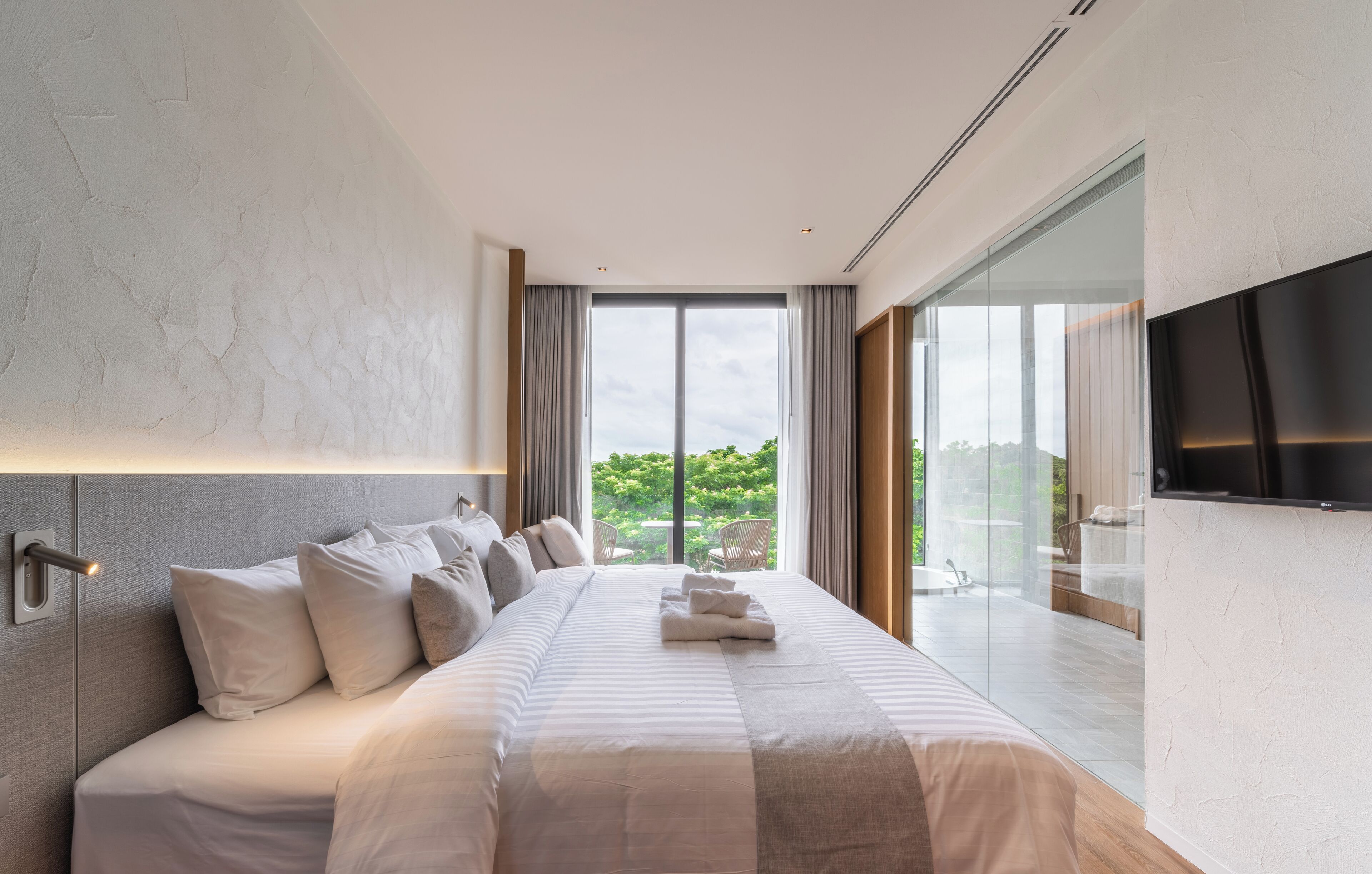 A modern bedroom with a large bed, a flat-screen TV, and a view of greenery outside.