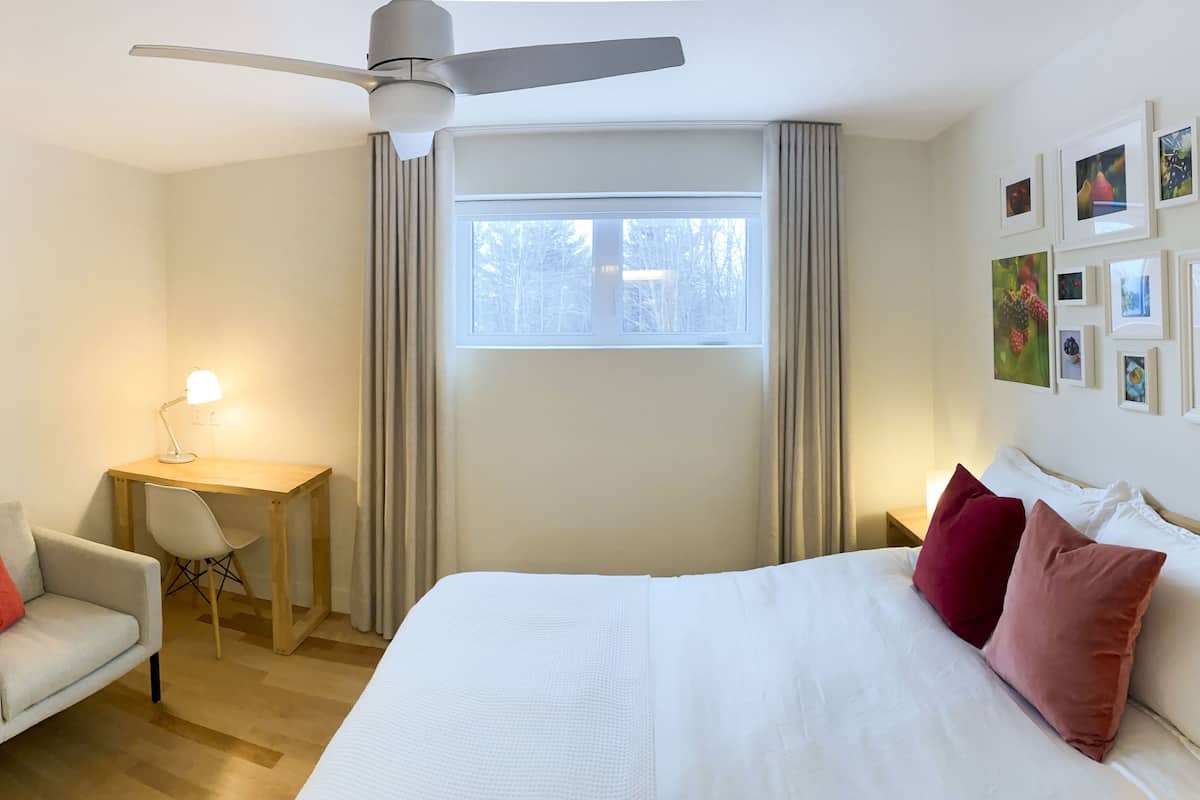 A hotel room with a bed, a desk with a chair, a ceiling fan, a window with curtains, and framed pictures on the wall.