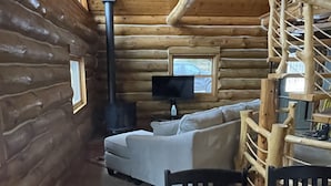 Een blokhut van binnen met een bank, een eettafel, stoelen, een open haard en een aan de muur bevestigde televisie.