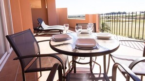 A round table with four chairs, a glass of water, and plates on a balcony with a view of a fence and greenery.