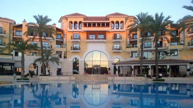 A hotel with a central pool reflecting the building and palm trees.