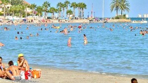 A crowded beach with people swimming and sunbathing, palm trees, and buildings in the background.