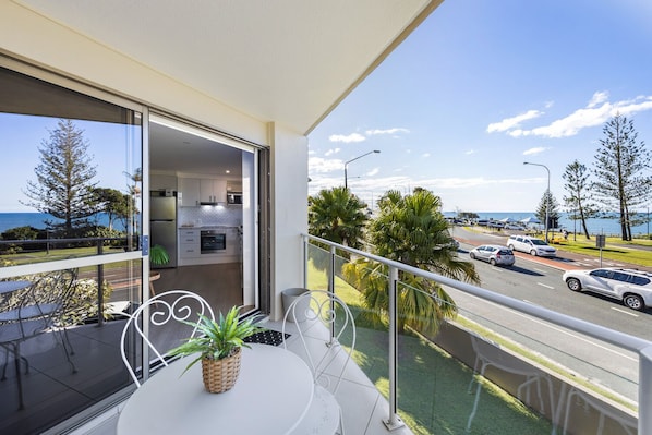 Varanda com vista para uma estrada e o mar, uma mesa de jantar com cadeiras e uma cozinha visível através de uma porta aberta.