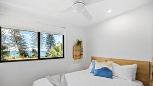 A bedroom with a large bed, a ceiling fan, a window with a view of trees and the sea, and a wall-mounted plant.