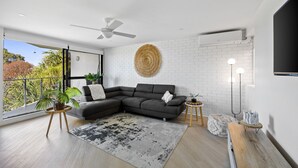 A modern living room with a grey sectional sofa, a round woven wall hanging, a large window with a view of trees, and a ceiling fan.