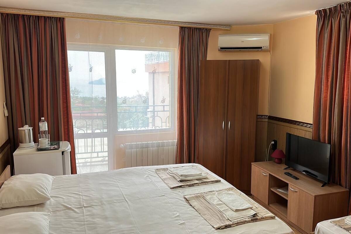A hotel room with a bed, a TV, a minibar, and a balcony with curtains.