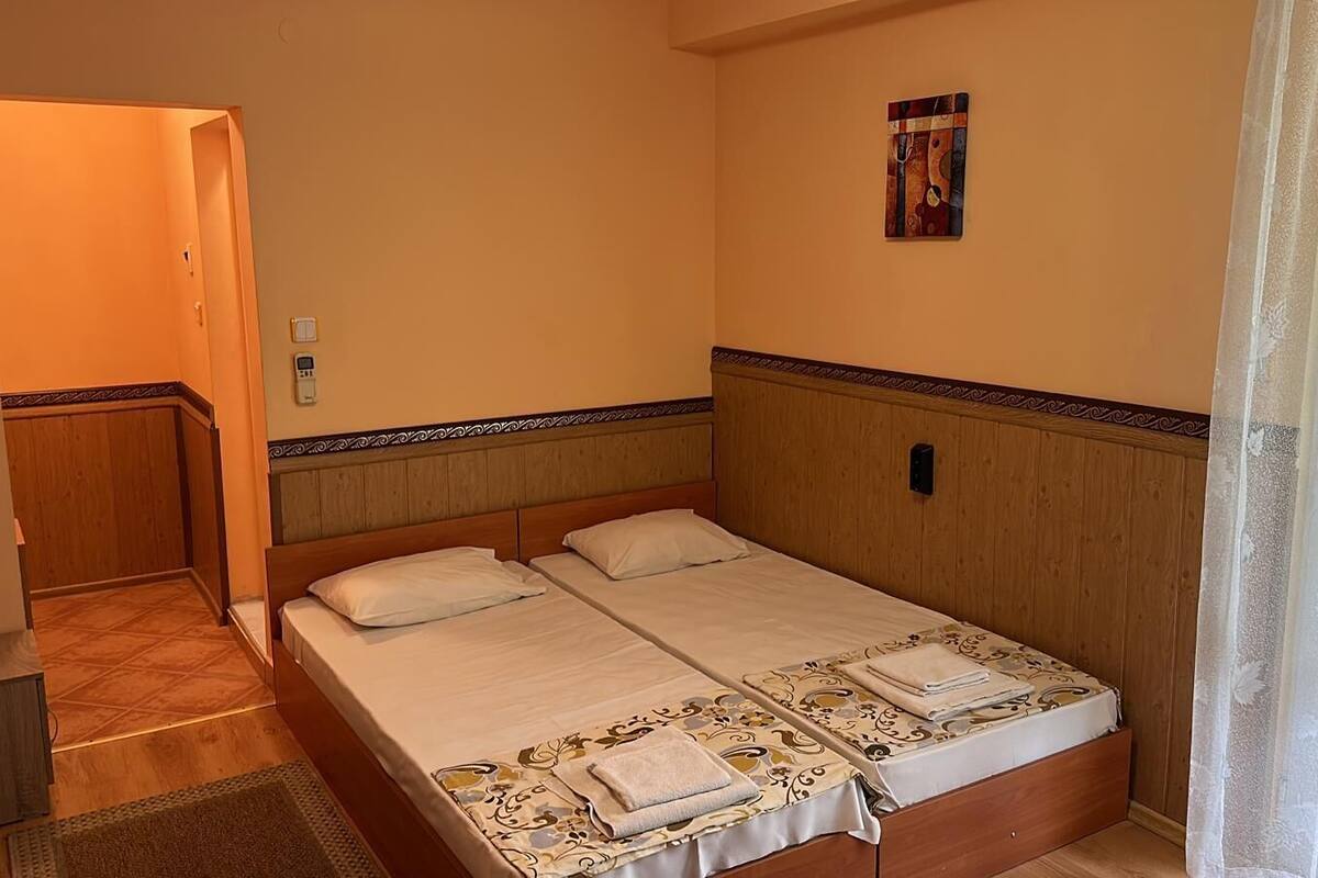 A hotel room with two beds, a wooden headboard, a small bedside table, and a window with sheer curtains.