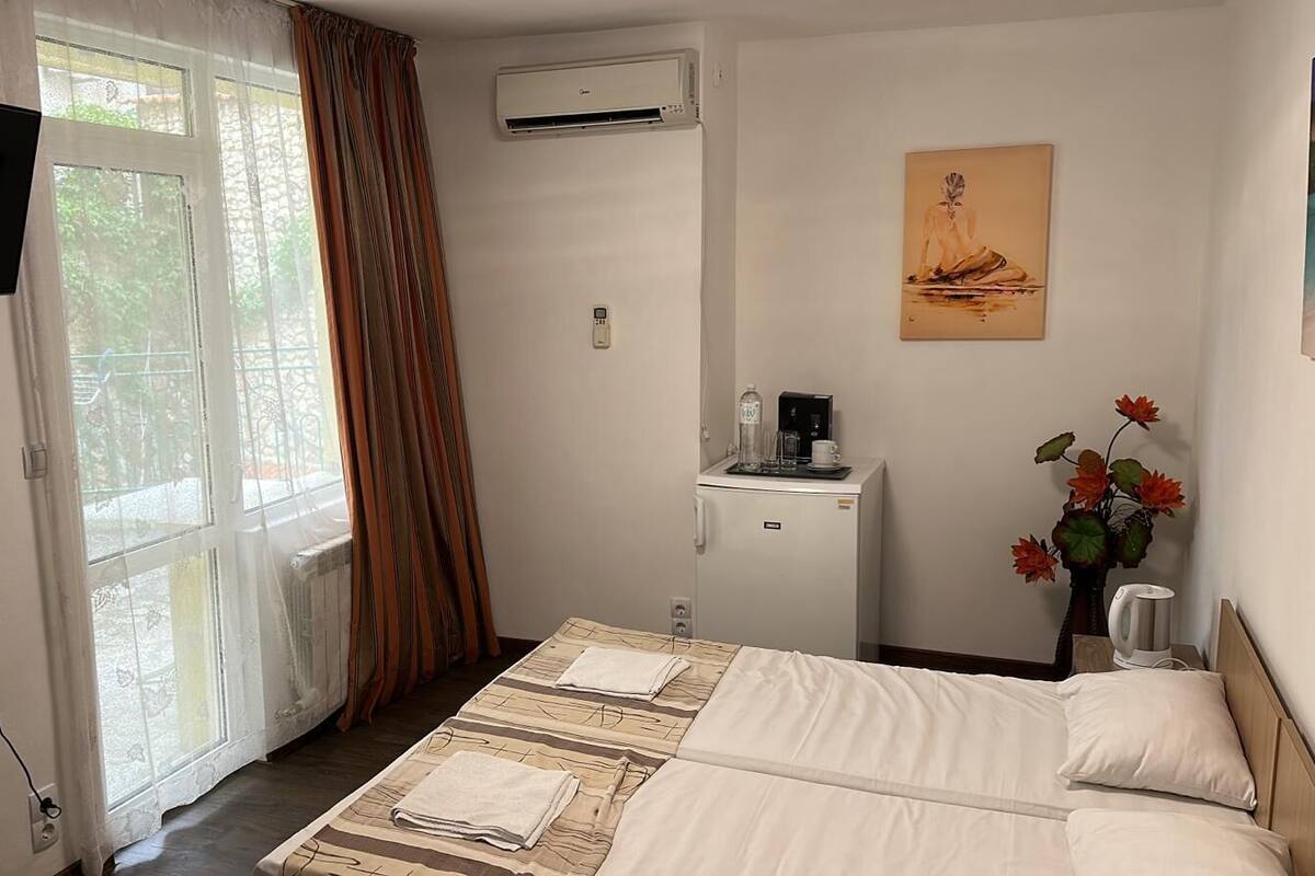 A hotel room with a bed, a desk with a coffee maker, an air conditioner, and a painting on the wall.