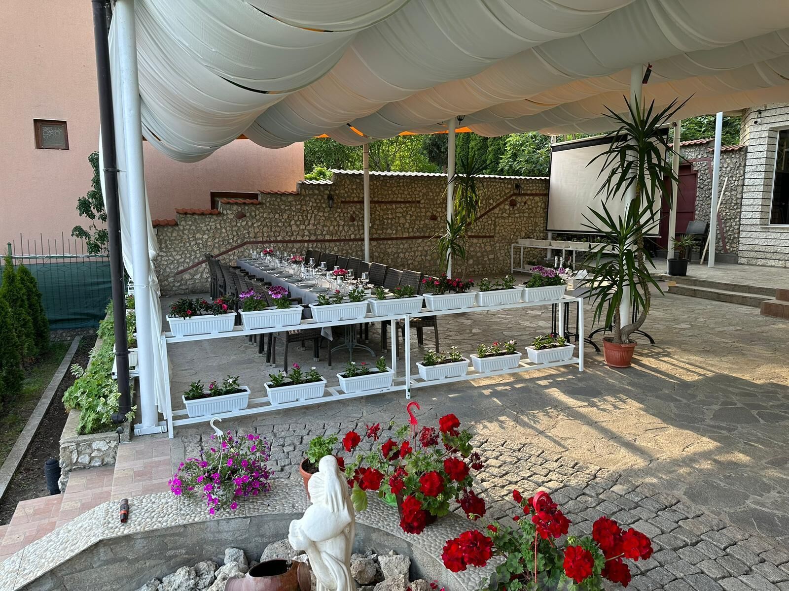 Un espace extérieur aménagé pour les repas, avec des tables et des chaises blanches, entouré de plantes et de fleurs en pot.