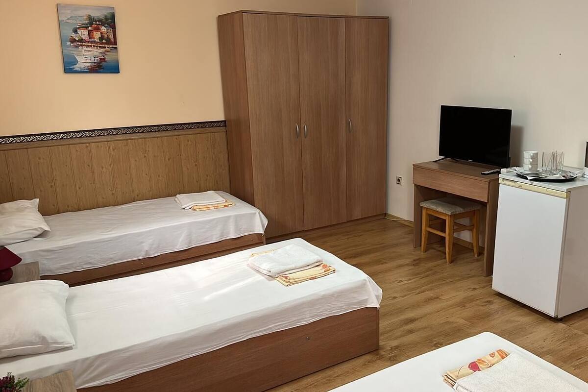 A hotel room with two single beds, a wooden wardrobe, a desk with a TV, and a small kitchenette with a refrigerator and coffee maker.