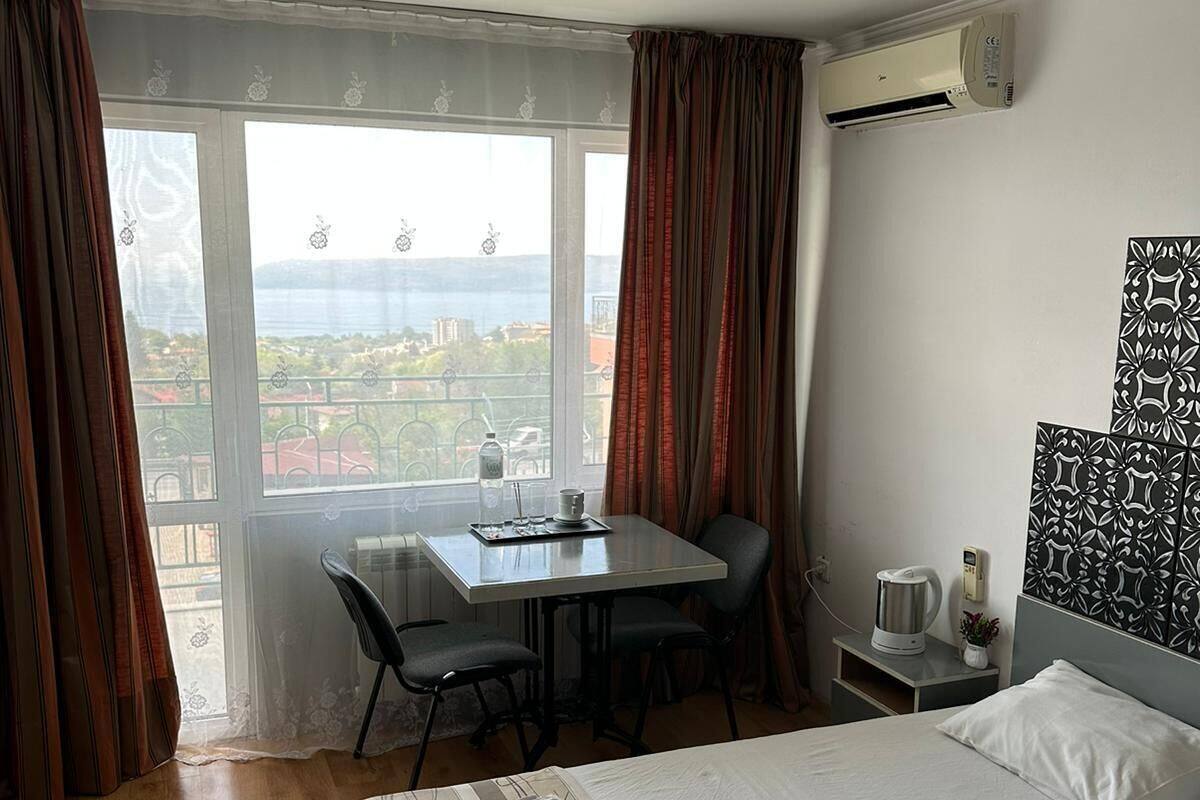 A hotel room with a large bed, a desk with chairs, a view of the city, and an air conditioning unit.
