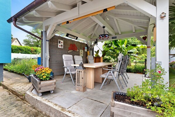 A covered patio with a table and chairs, a wooden planter with flowers, and a lantern.