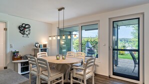 Een eetgedeelte met een ronde tafel, witte stoelen, een hanglamp en uitzicht door grote ramen.