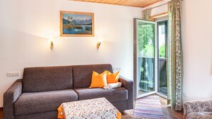 Una habitación con sofá, una mesa con mantel floral, un cuadro de un paisaje montañoso y una puerta corrediza de vidrio al exterior.
