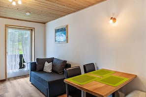 Una stanza con soffitto in legno, un divano, un tavolo con tovagliette verdi, un quadro appeso al muro e una finestra con tende leggere.