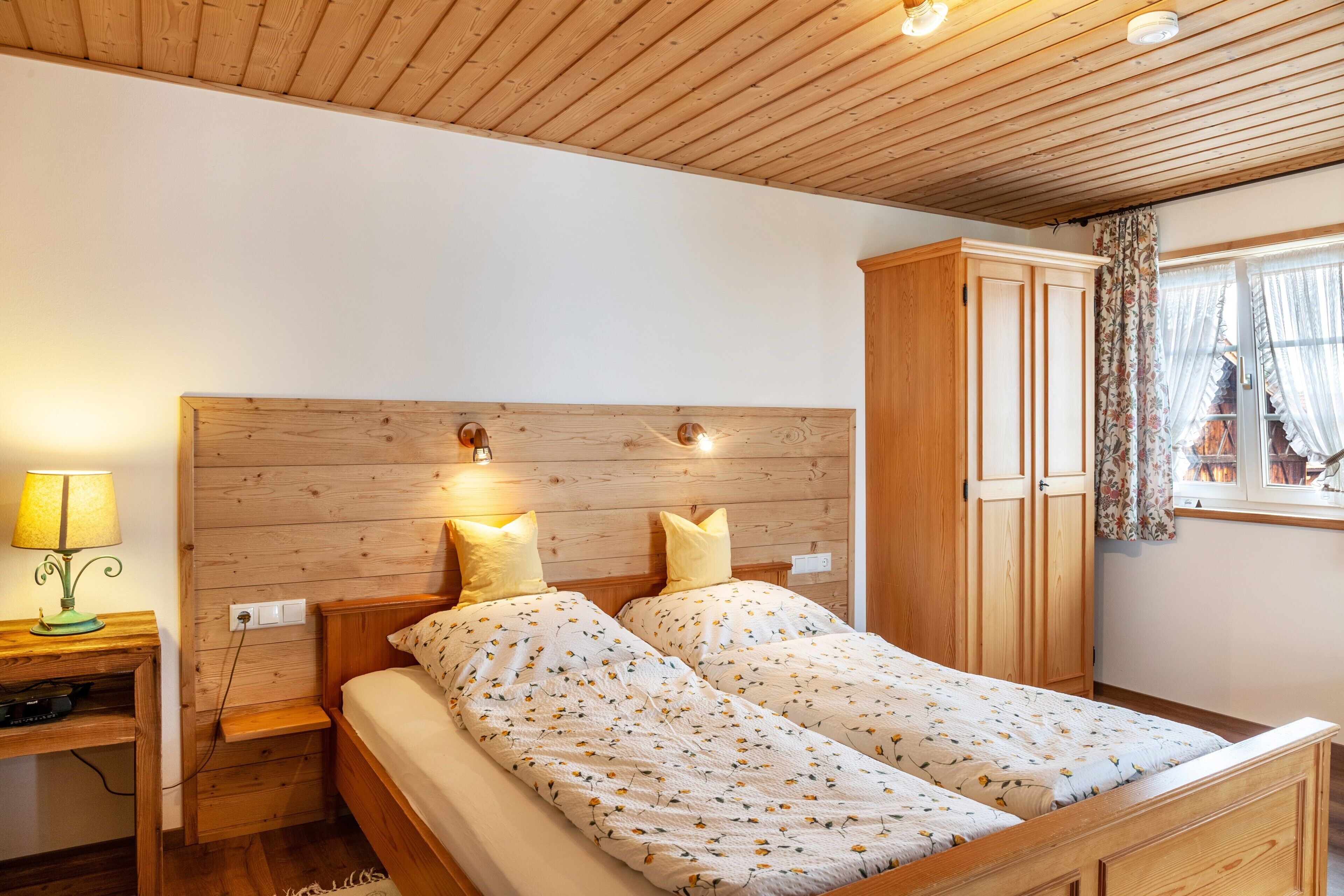 A bedroom with two beds, wooden headboard, bedside lamps, a wardrobe, and a window with curtains.