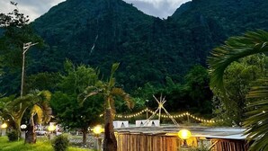 A paved pathway leading to a wooden structure with string lights, surrounded by lush greenery and palm trees, with a mountain in the background. - Xaymone Riverside Resort and Restaurant (Vang Vieng)