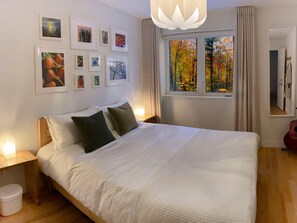 Ein Schlafzimmer mit einem großen Bett, vielen Kissen, einem Nachttisch mit Lampe, einem Fenster mit Blick auf Herbstlaub und mehreren gerahmten Bildern an der Wand.
