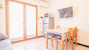 Une petite pièce lumineuse dotée d’une porte-fenêtre coulissante, d’un réfrigérateur, d’un four à micro-ondes, d’une télévision et d’une table à manger avec des chaises.