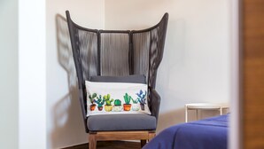 Egy szobában található szék, melynek a párnáján kaktuszminta látható, a padló fából készült, és egy kis fehér asztal áll benne, a falak pedig egyszerűek.