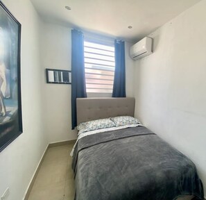 Une petite chambre à coucher, bien rangée, avec un lit simple, une fenêtre avec des rideaux, un appareil de climatisation et un tableau encadré au mur.