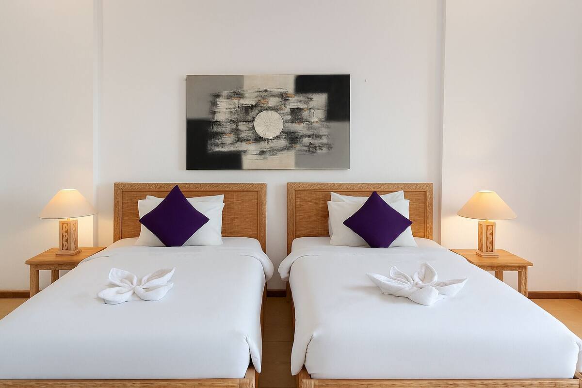 Quarto de hotel com duas camas, cada uma com cabeceira de madeira, roupa de cama branca e almofadas roxas. Há um quadro na parede acima das camas e duas luminárias de cabeceira em mesas de cabeceira de madeira.