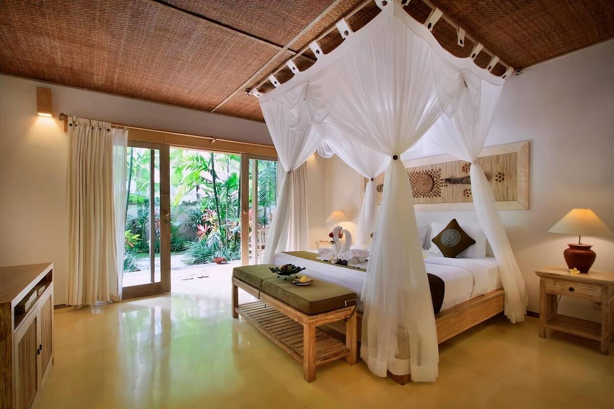 Quarto com cama, um banquinho, mesa de cabeceira e vista para o verde lá fora.
