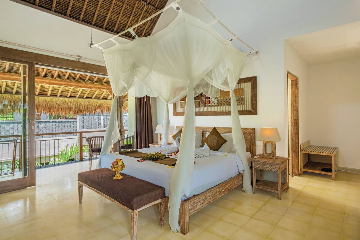 Um quarto espaçoso com cama de dossel, um banco de madeira, uma mesa de cabeceira e vista para uma construção com telhado de palha.