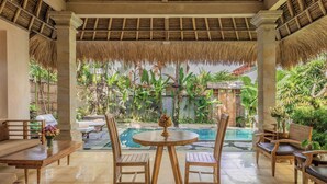 Zona junto a la piscina con muebles de madera, una mesa redonda con fruteros y vistas a una piscina y abundante vegetación.