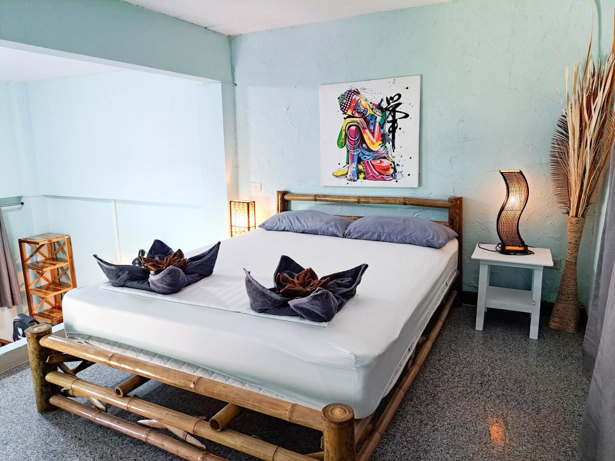 A bed with a wooden frame, two pillows, a bedside table with a lamp, and a wall with a colorful abstract painting.