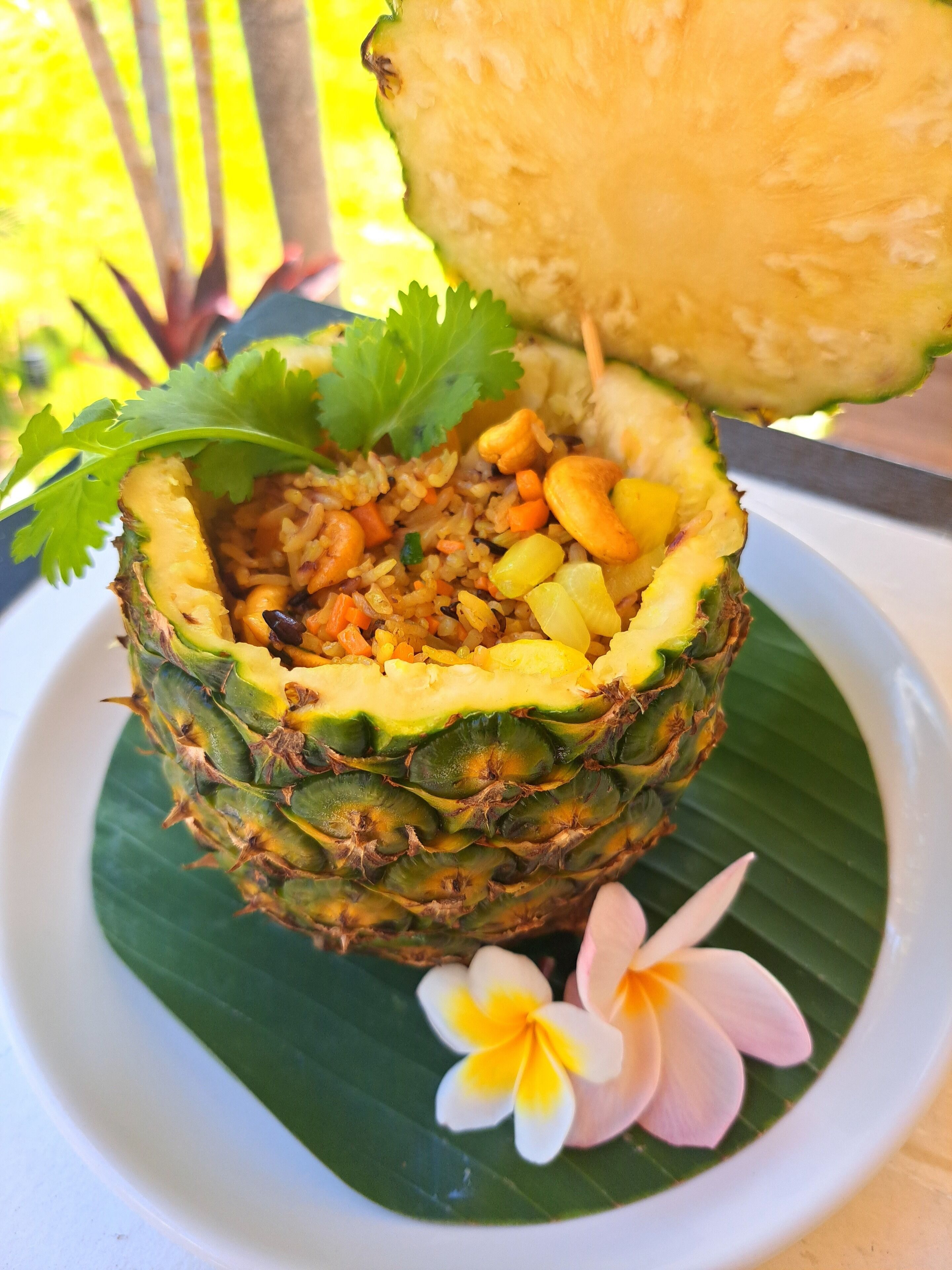 Un plato servido en una cáscara de piña con arroz, verduras y nueces, decorado con una hoja de cilantro y una flor.