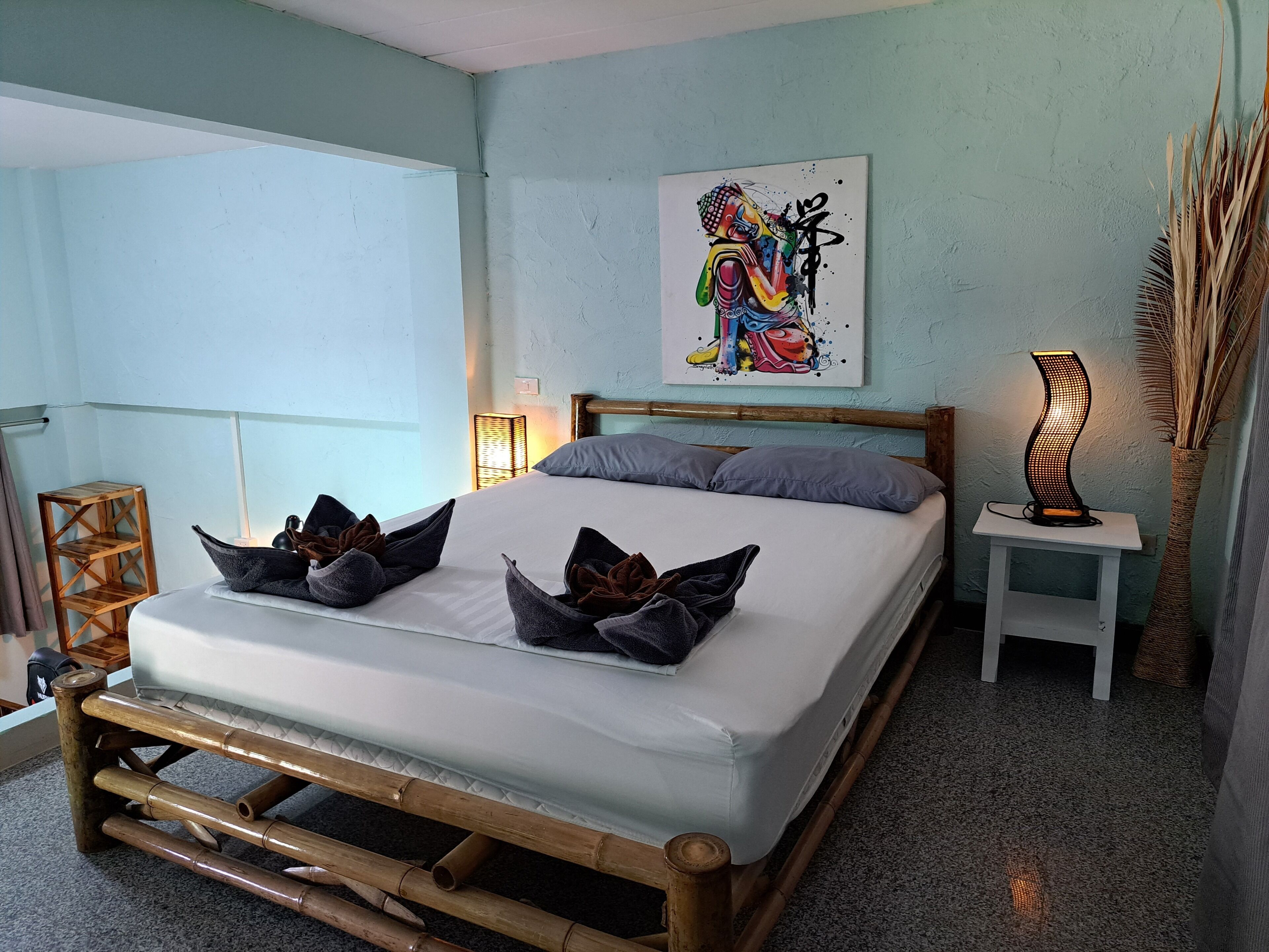 A bed with a bamboo frame, grey pillows, and a colorful abstract painting on the wall.