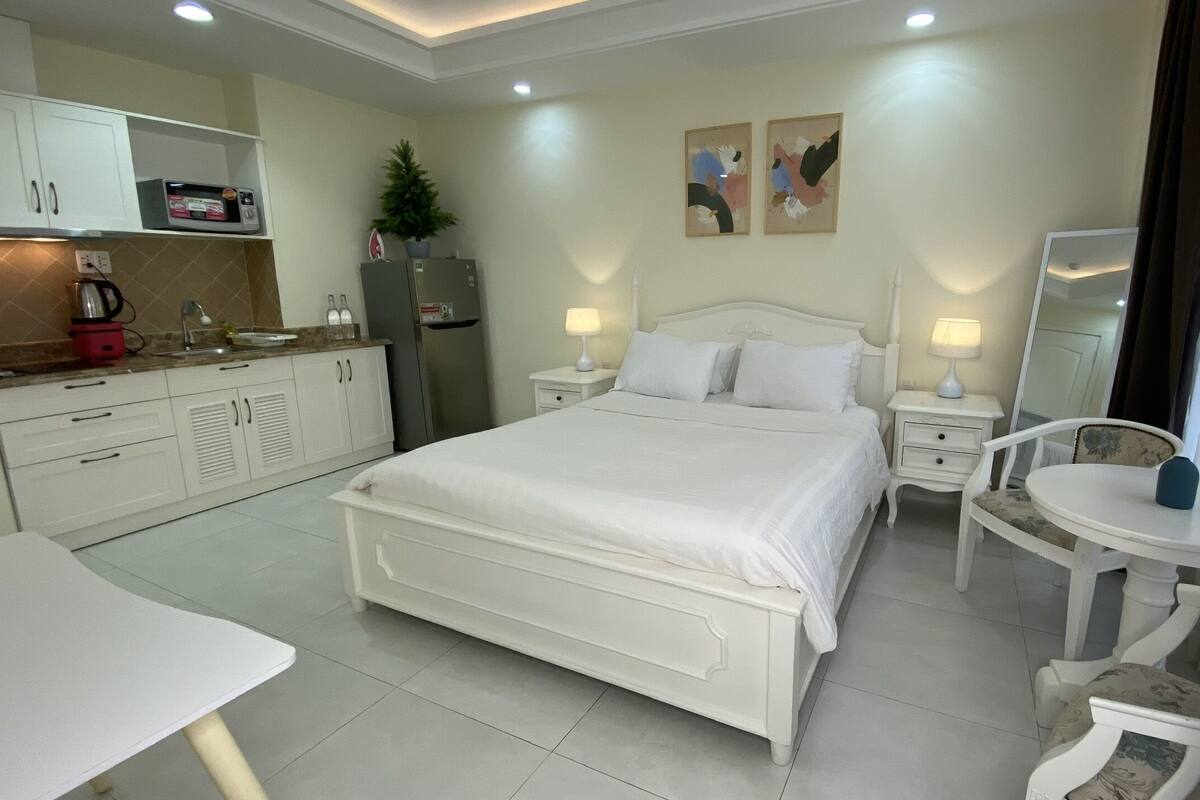 Une chambre d’hôtel comprenant un lit, une petite table, une chaise, un miroir, une table de chevet, un réfrigérateur, un four à micro-ondes, un lavabo et une plante en pot.