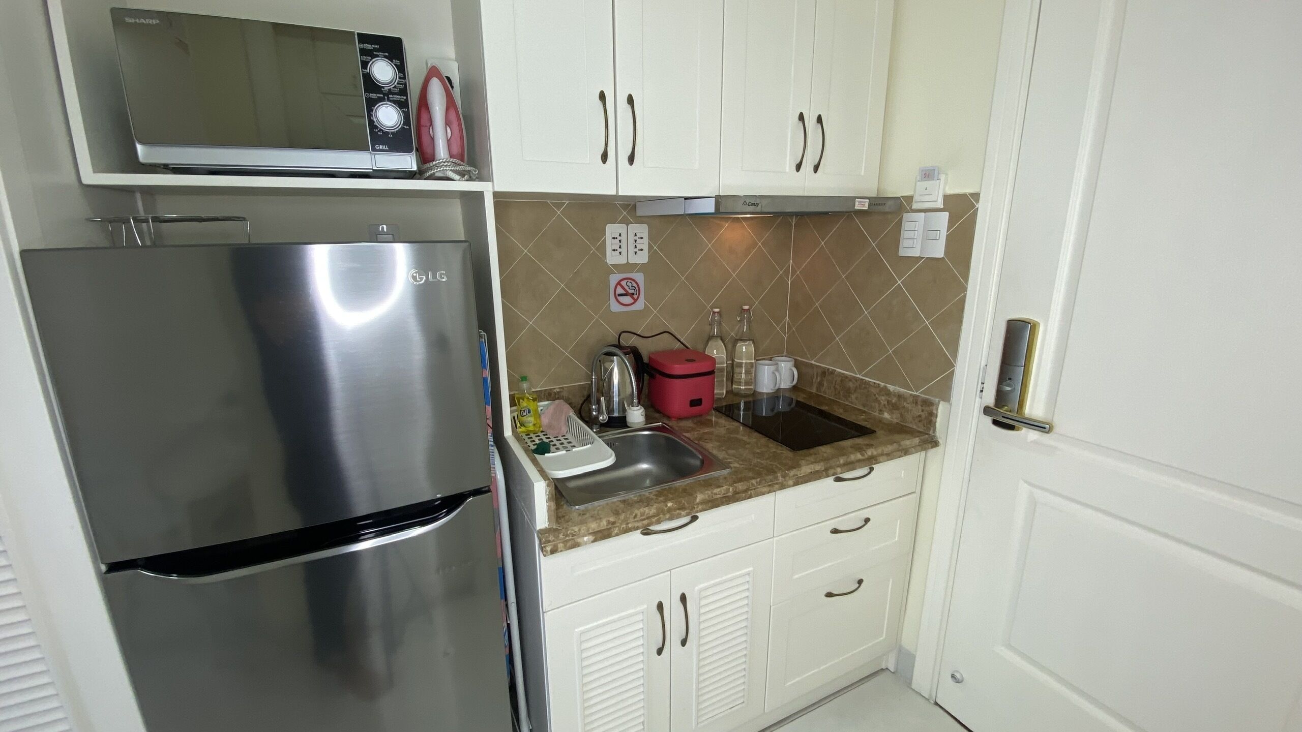 A compact kitchen with a stainless steel refrigerator, a microwave, a kettle, and a sink.